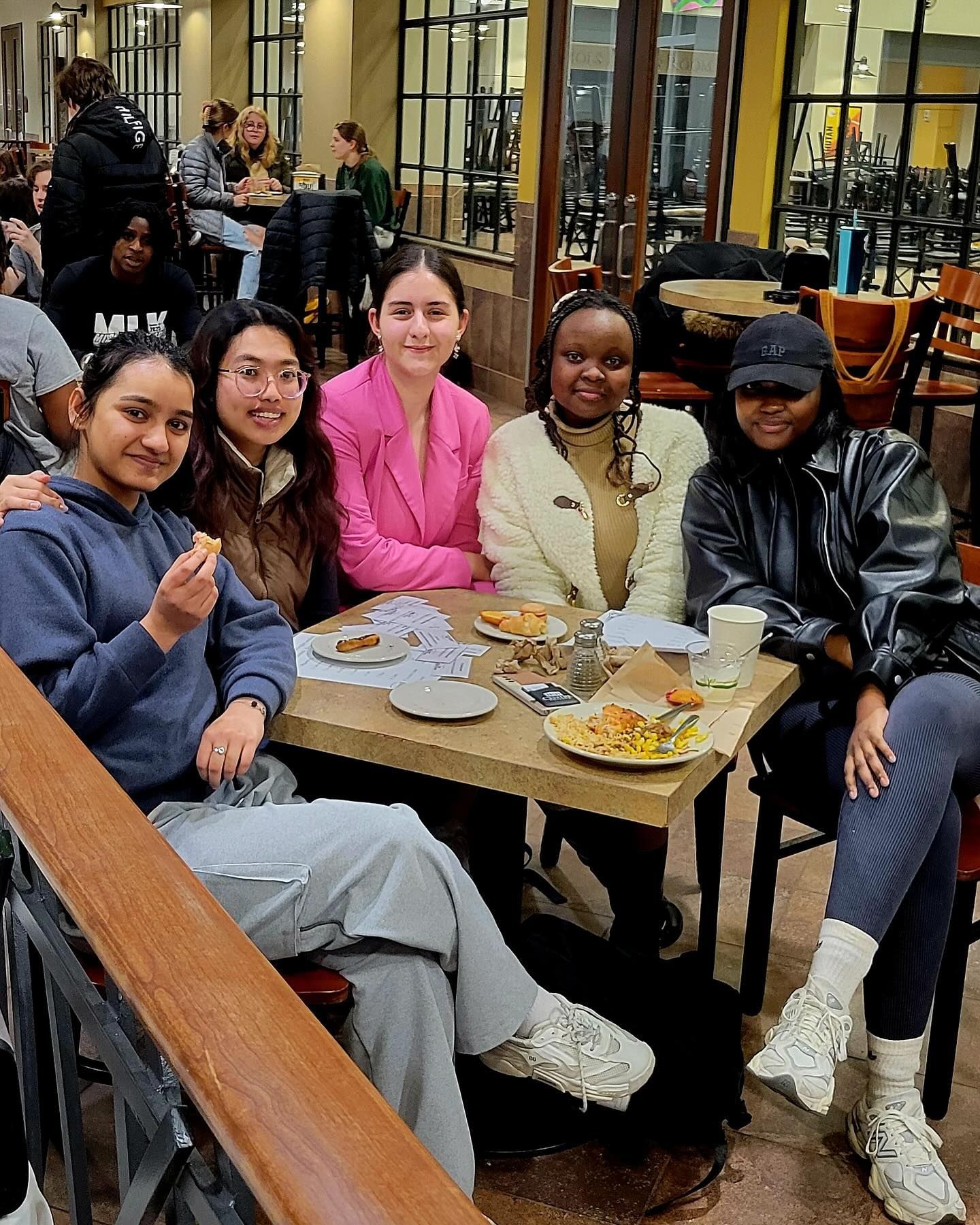 Five people sit around a wooden table in a cafe, talking and eating, with plates of food.