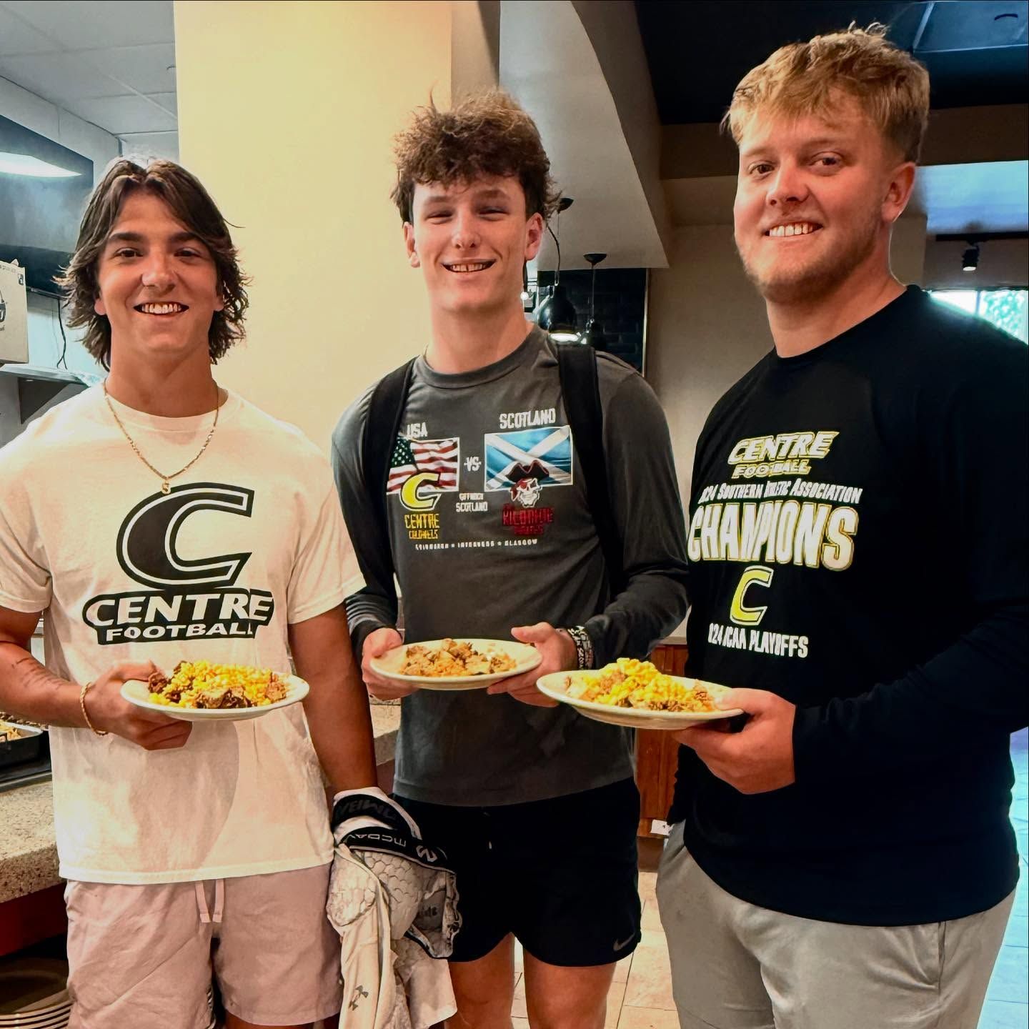 Three students in Centre College gear stand smiling, each holding a plate of food.