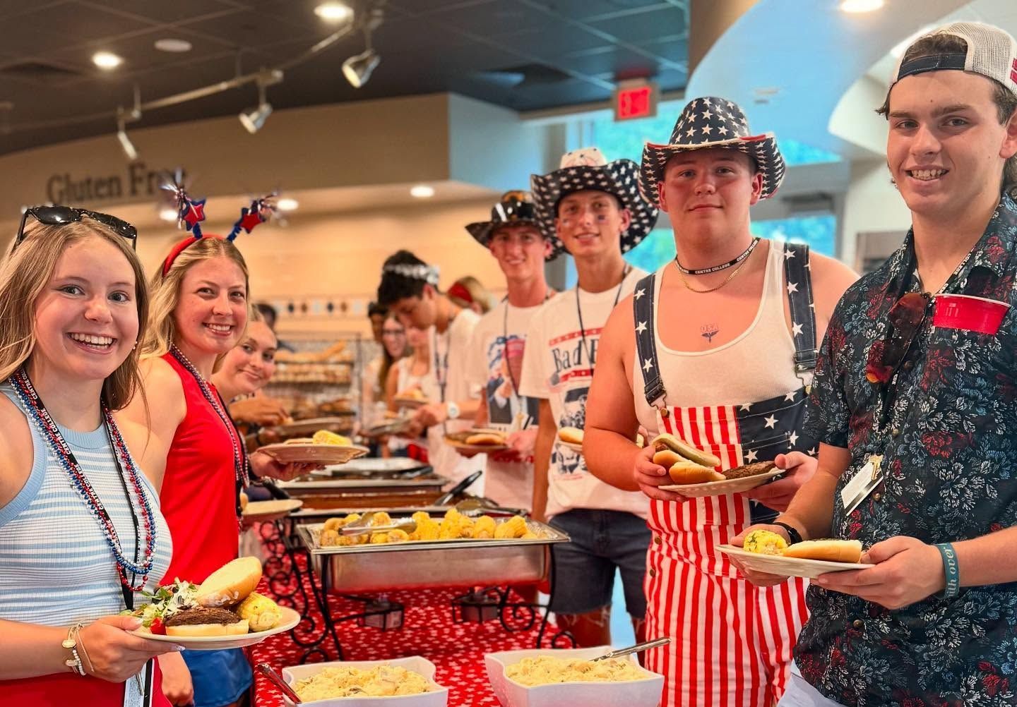 A group of people wearing patriotic-themed hats and clothing smile while gathering food at a buffet line.
