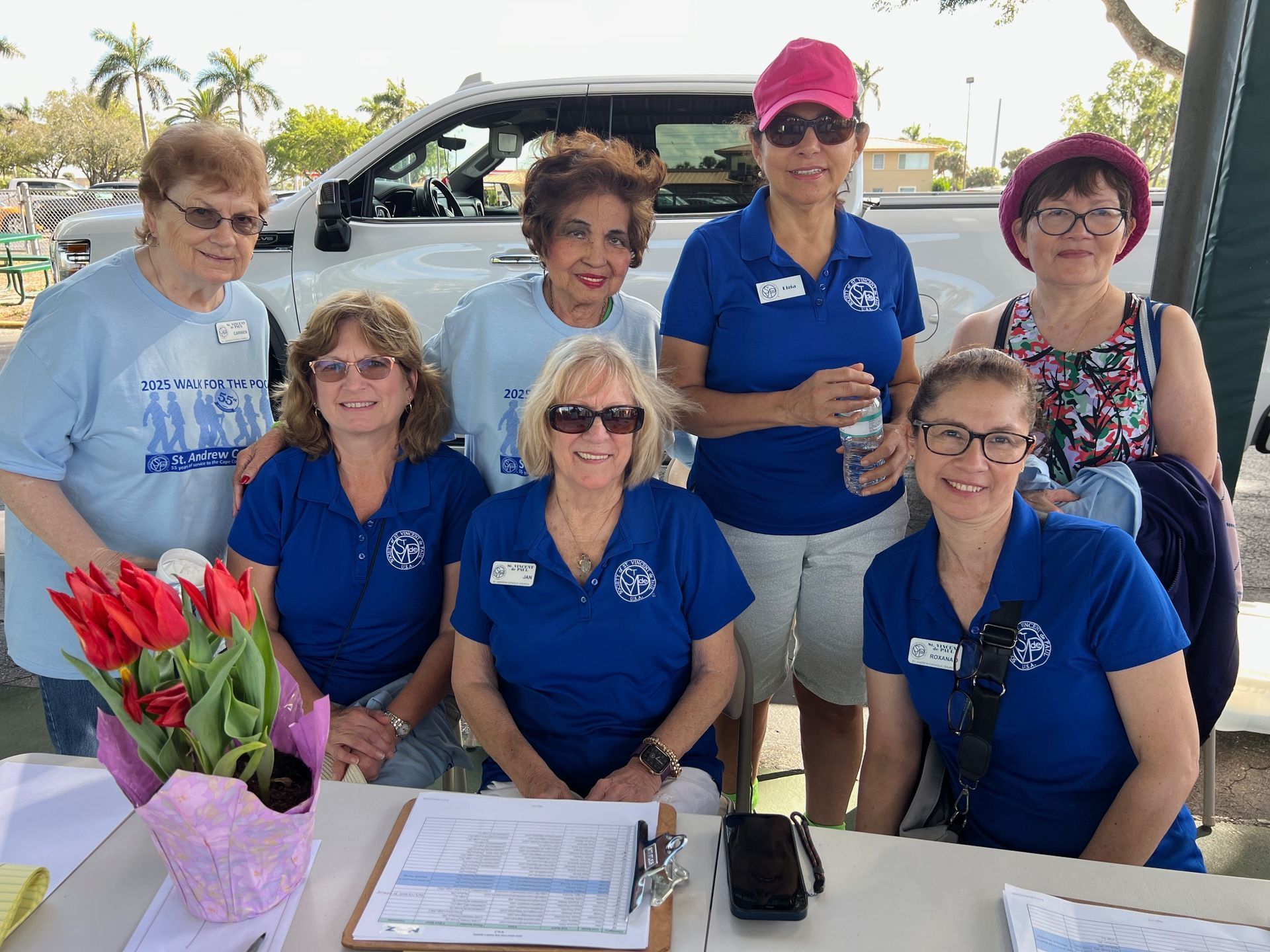 Group of volunteers at a table, wearing blue shirts. They are outdoors with a truck and flowers visible.