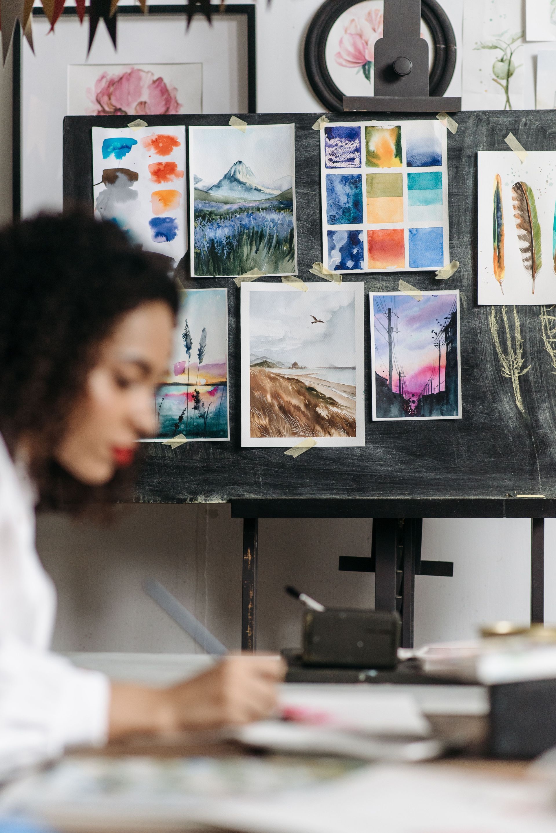 Artist painting at a table in front of a board of colorful landscape sketches and color swatches
