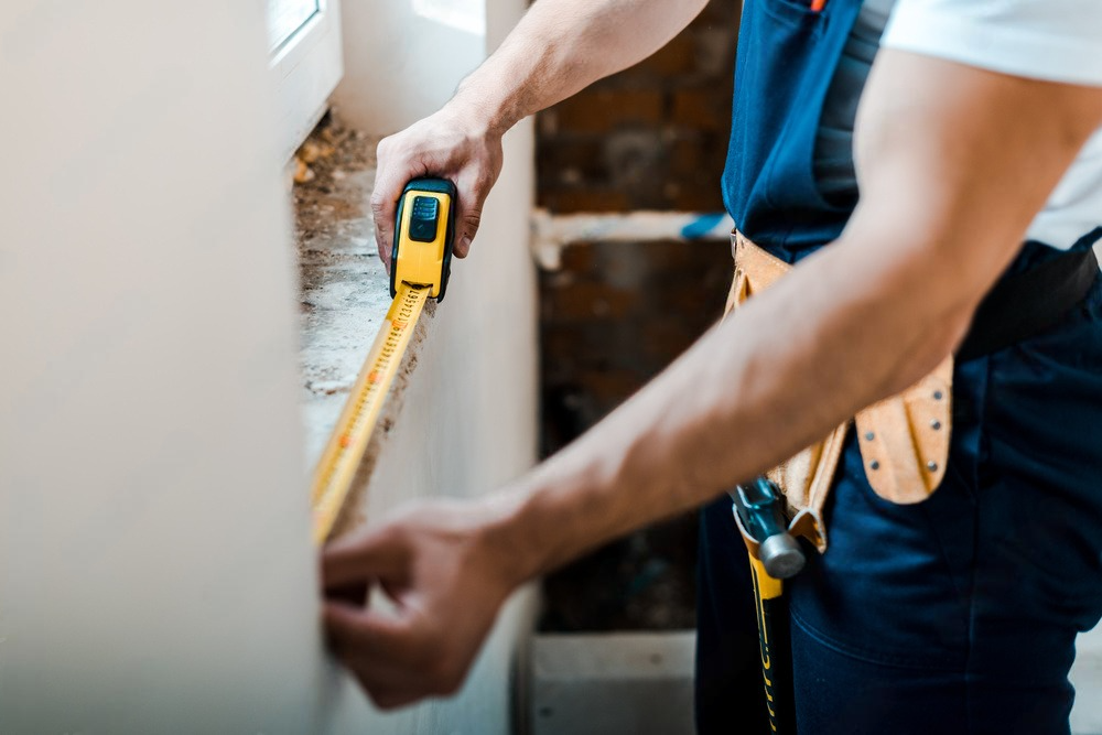 Person in blue overalls measuring a wall with a yellow tape measure.