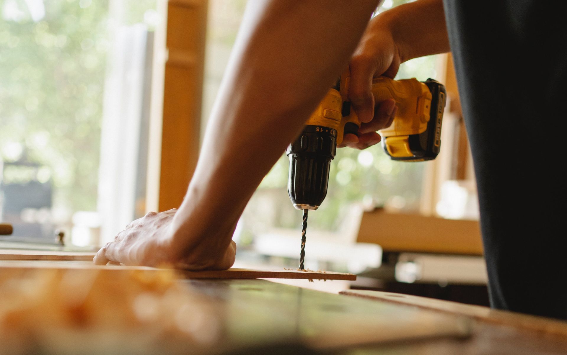 Person using a yellow drill to bore a hole into a wooden plank at a workshop, near a window.