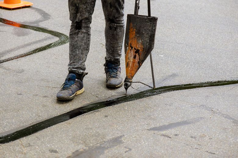 A person laying down a dark green line on pavement using a handheld applicator.