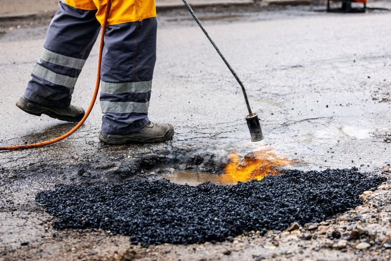 Worker in safety gear using a flame torch to repair asphalt road surface.