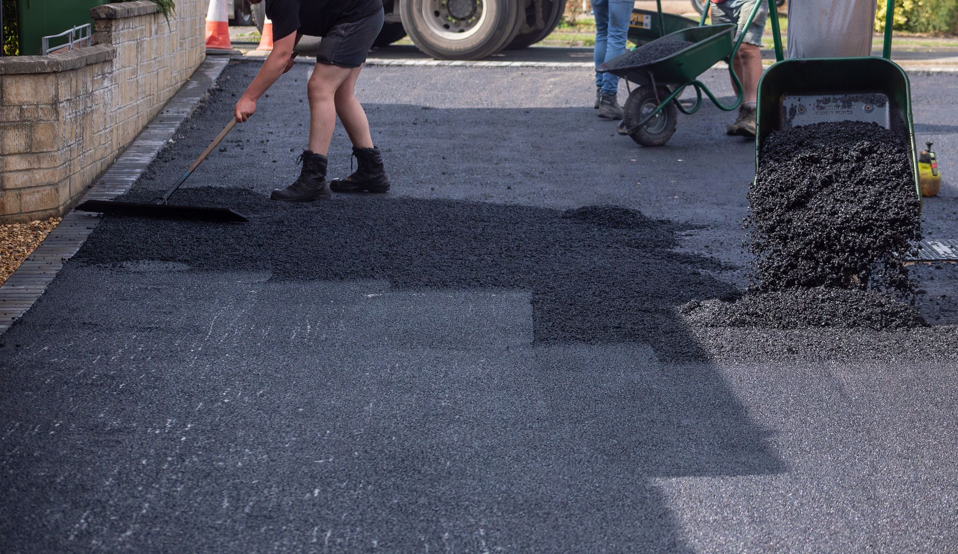 Man raking fresh asphalt on a driveway next to a brick wall. A truck and wheelbarrow are present.