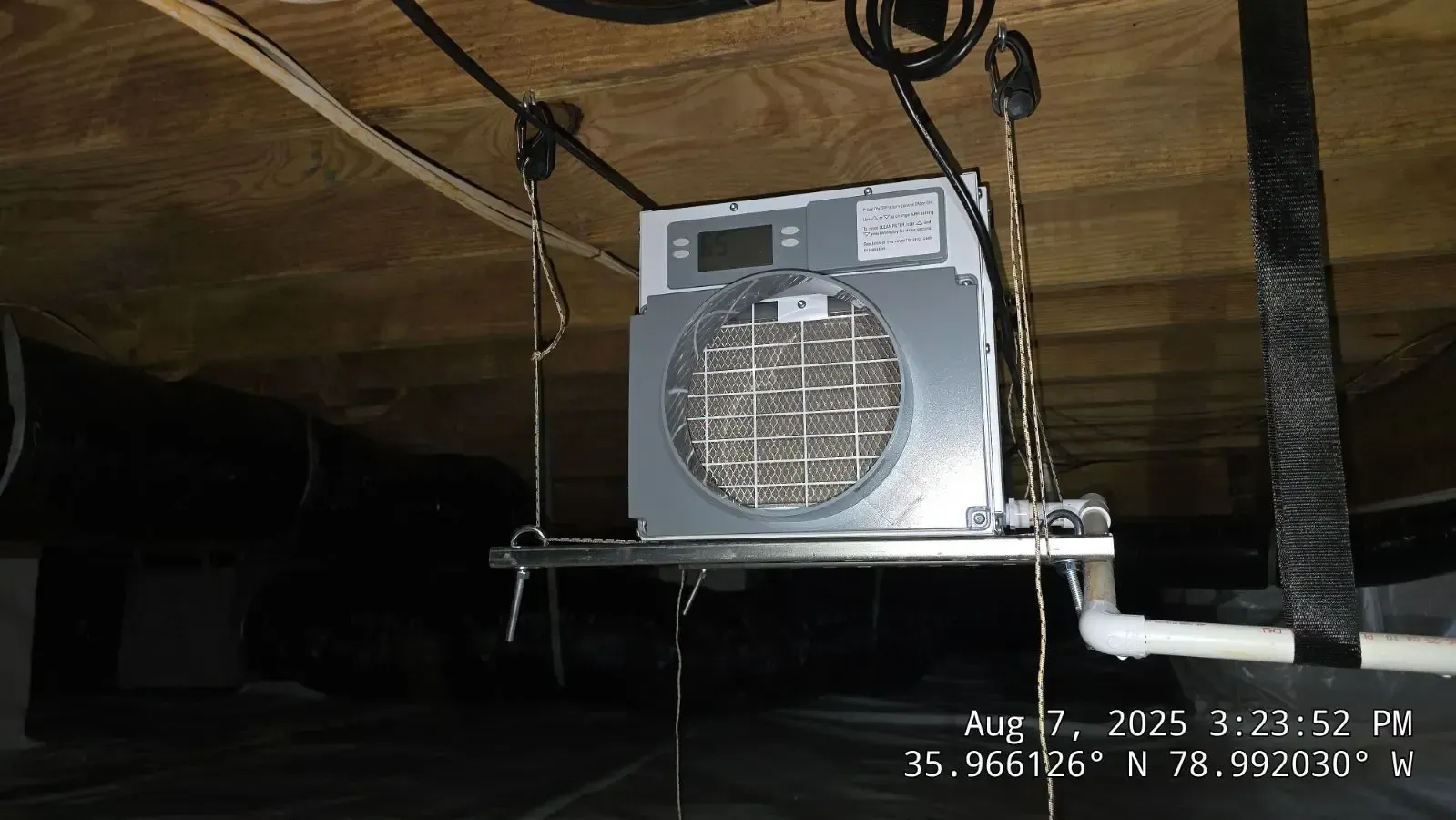 Silver dehumidifier suspended in a crawl space, with plumbing and electrical wiring visible.