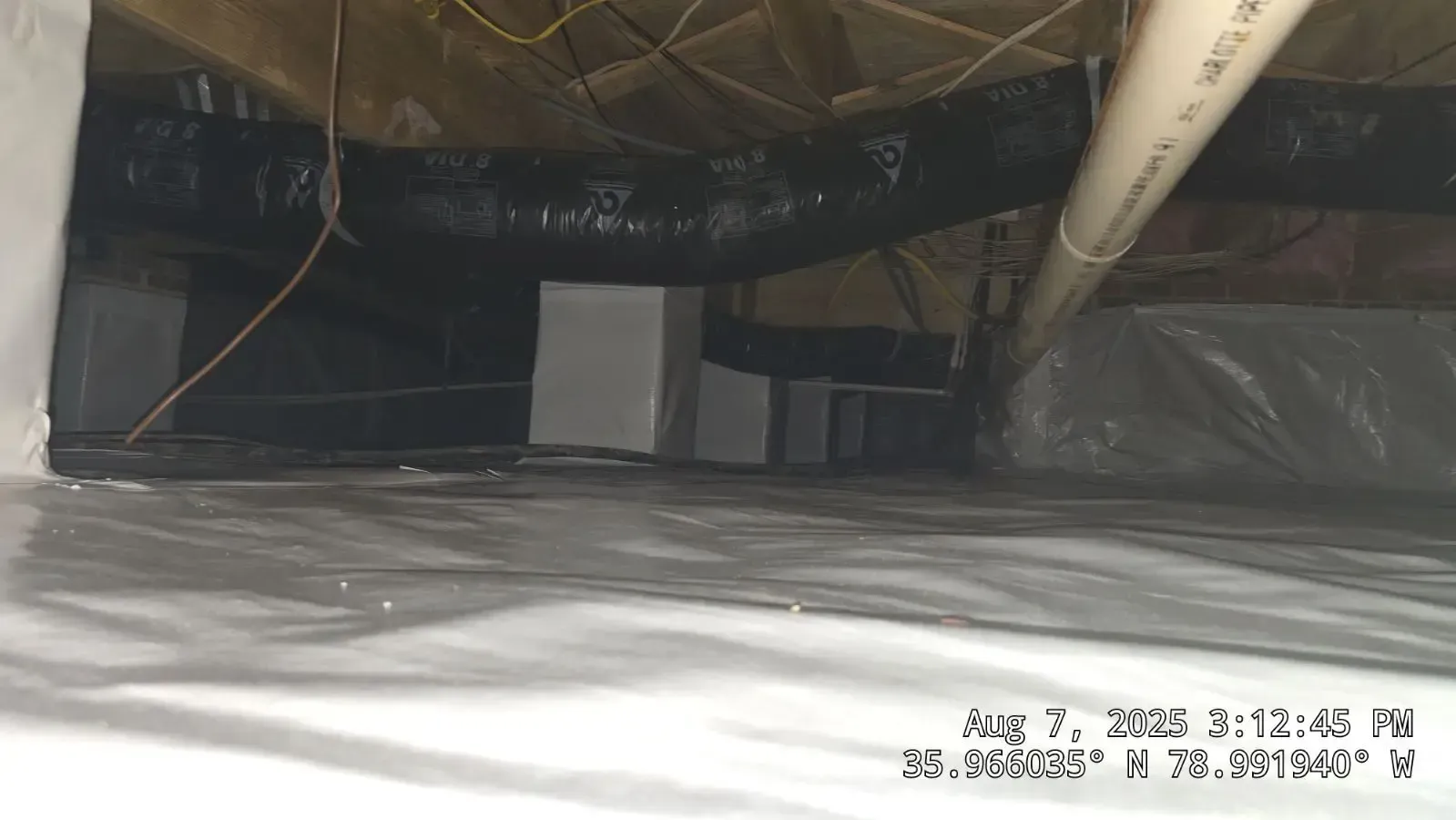 Crawlspace with insulation and ductwork. White blocks support beams, beneath a house.