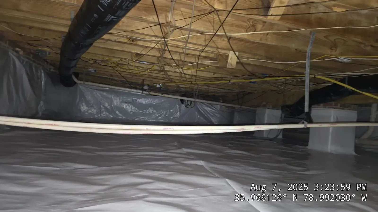 Crawlspace with white vapor barrier on floor, wooden beams, black flexible ducting, and support pillars.