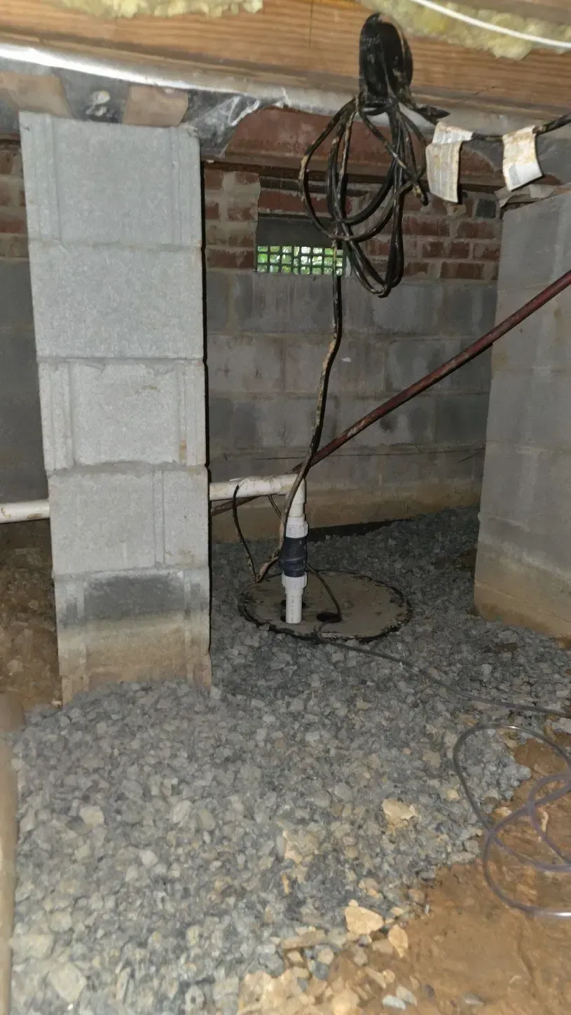 Sump pump in a crawlspace. Gray cinder block columns and walls, gravel floor. Cables overhead.