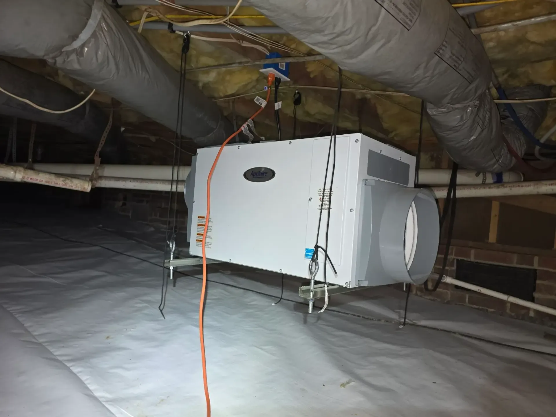 A large, white HVAC unit suspended in a crawlspace, with ductwork and wiring visible.