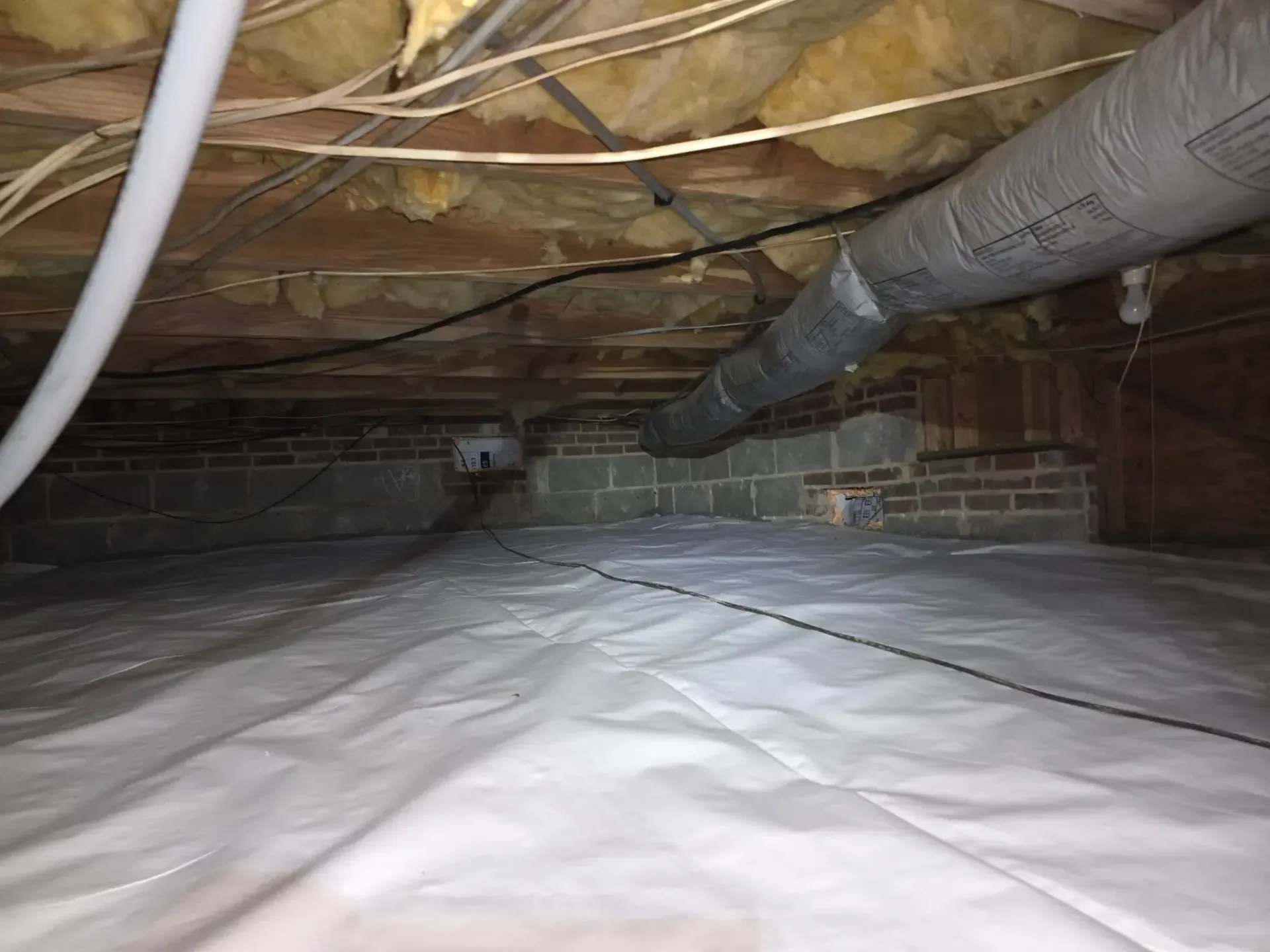 Crawl space with white vapor barrier, brick foundation, ductwork, and electrical wiring.