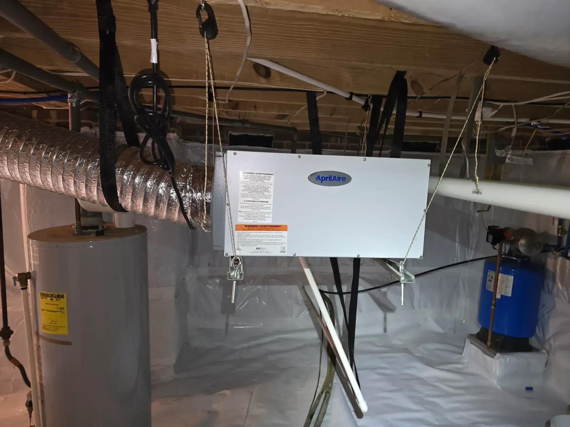 A crawl space with a water heater, HVAC unit, and water tank. White plastic sheeting covers the ground.