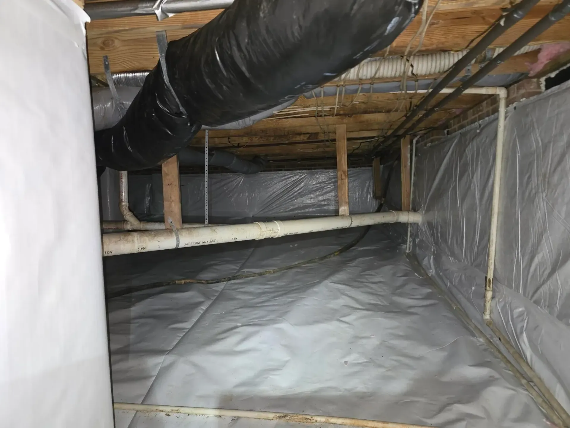Crawlspace with gray vapor barrier on floor and walls; plumbing and ductwork visible.