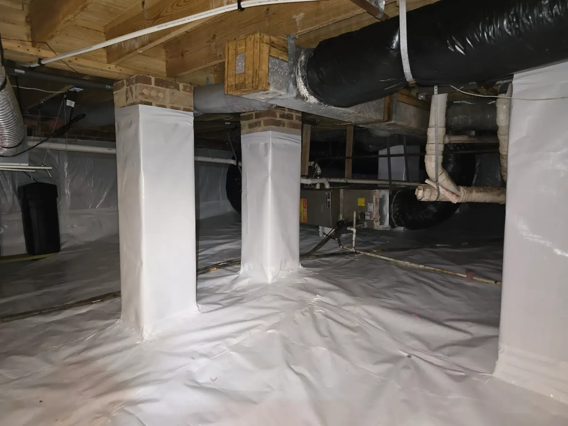 Crawl space with white vapor barrier on floor and columns wrapped in white plastic. Wooden supports and ductwork are visible.