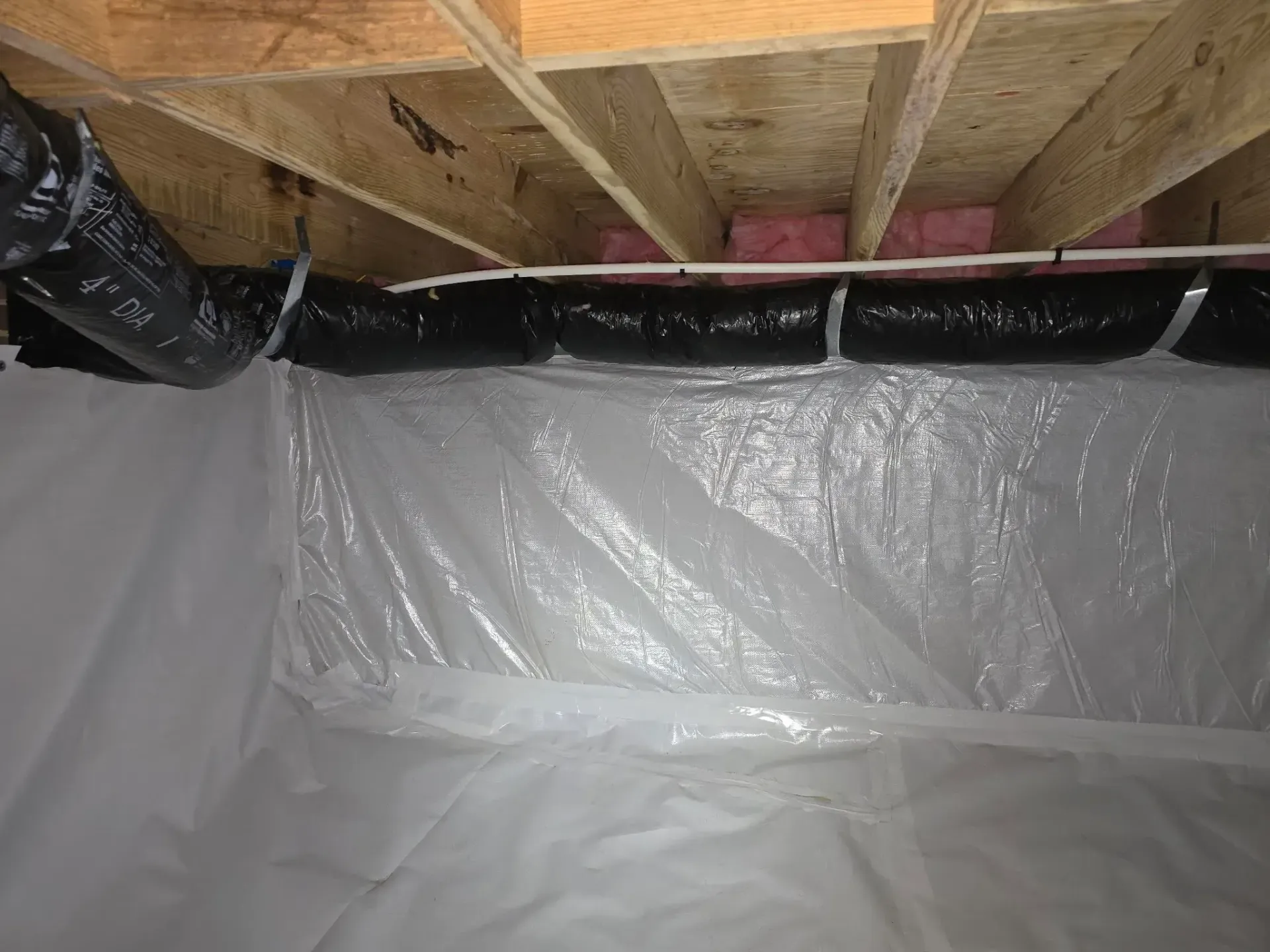 Crawlspace with plastic sheeting on walls and floor, insulation above, and black ductwork. Wooden beams overhead.