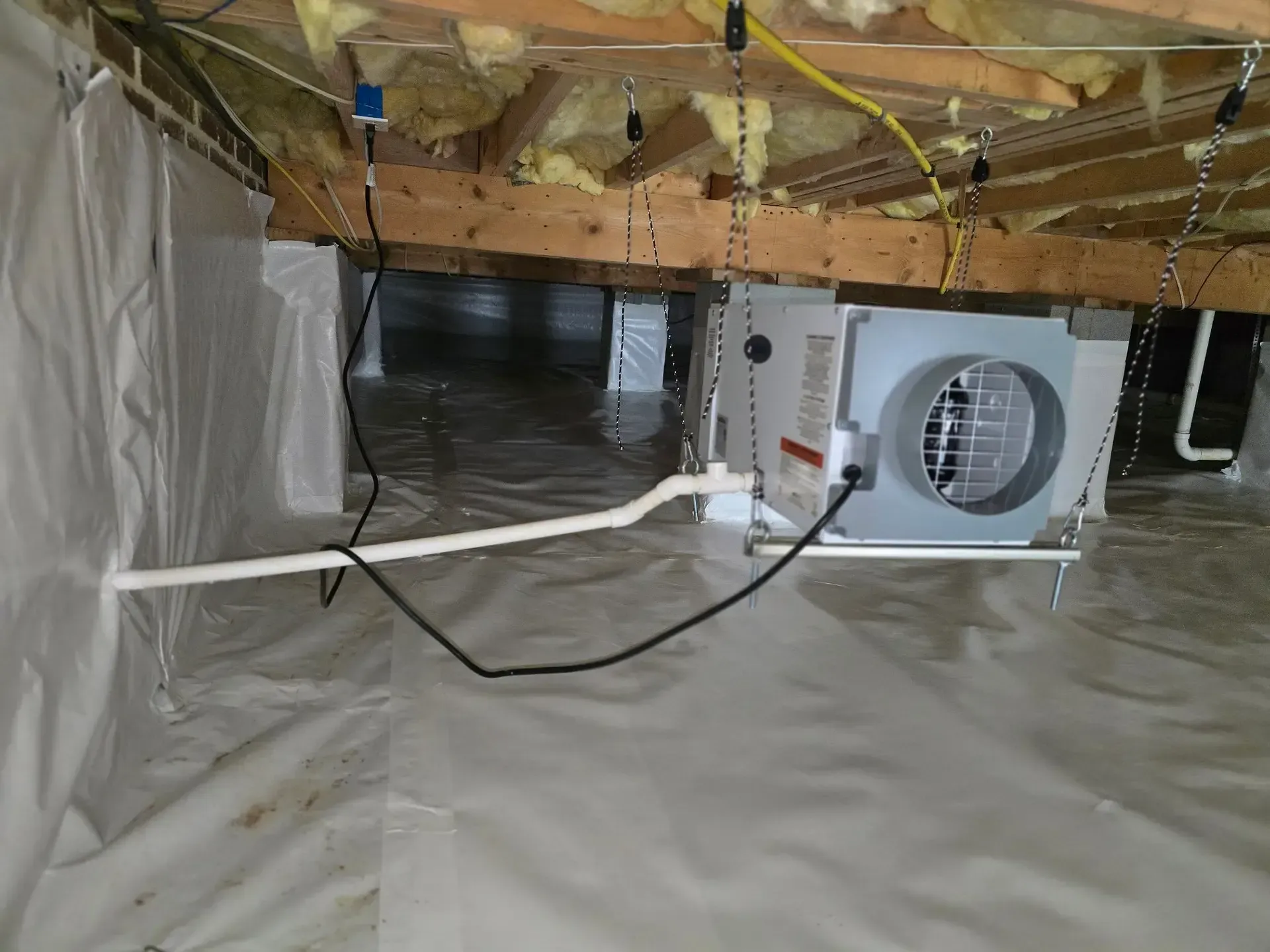 A crawl space with a white vapor barrier, wooden beams, and a dehumidifier.