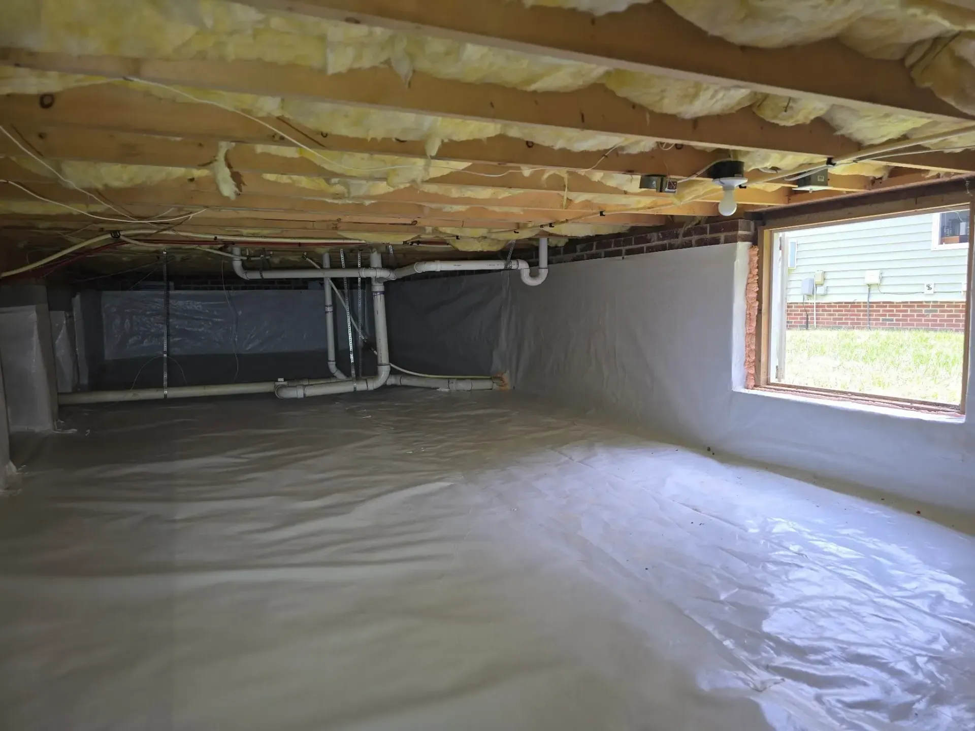 Crawlspace with white plastic sheeting on the ground and walls. Pipes and insulation are visible on the ceiling. Window on the right.