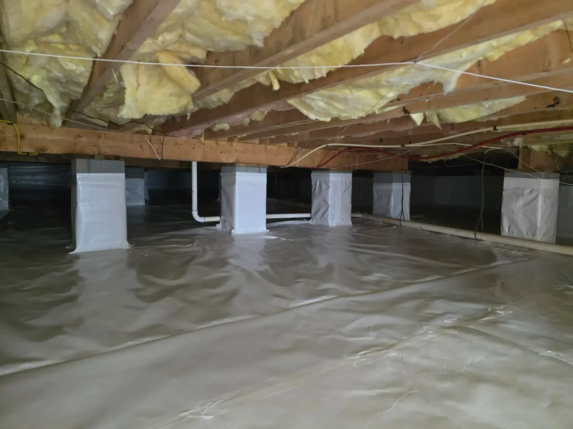 Crawl space under a house, with insulation, support columns, and plastic sheeting covering the ground.