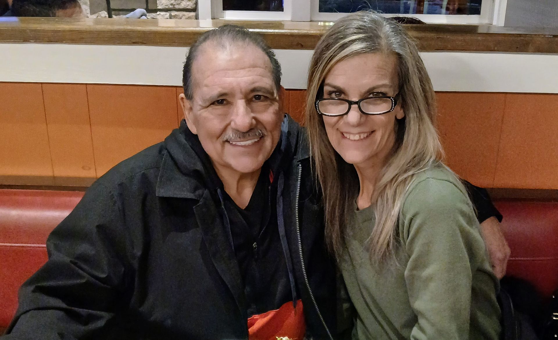 Man and woman smiling, sitting close together on a red booth. The man wears a black jacket, the woman wears glasses and green shirt.