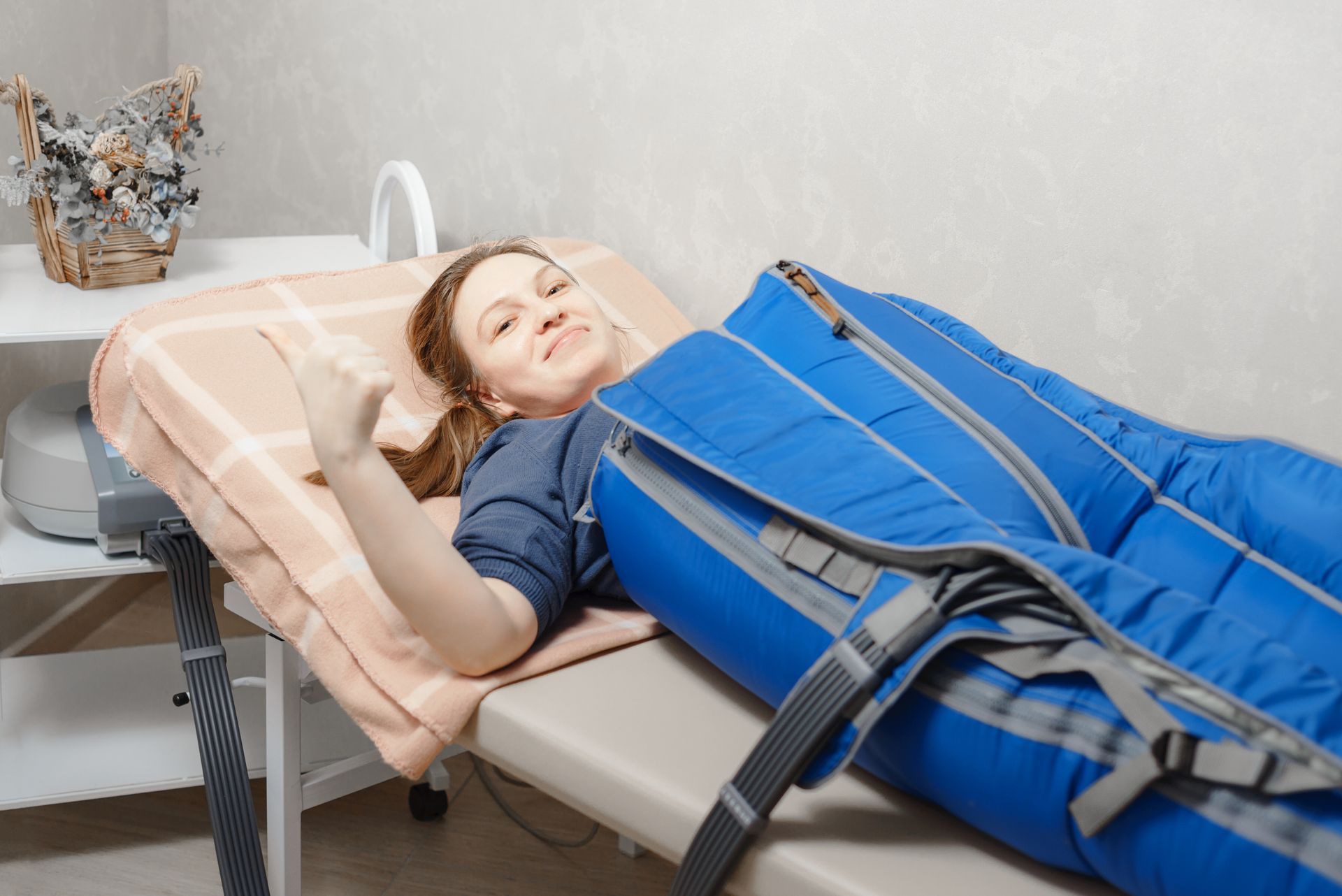 A person lies on a treatment table with blue compression boots on their legs, smiling and raising a fist.