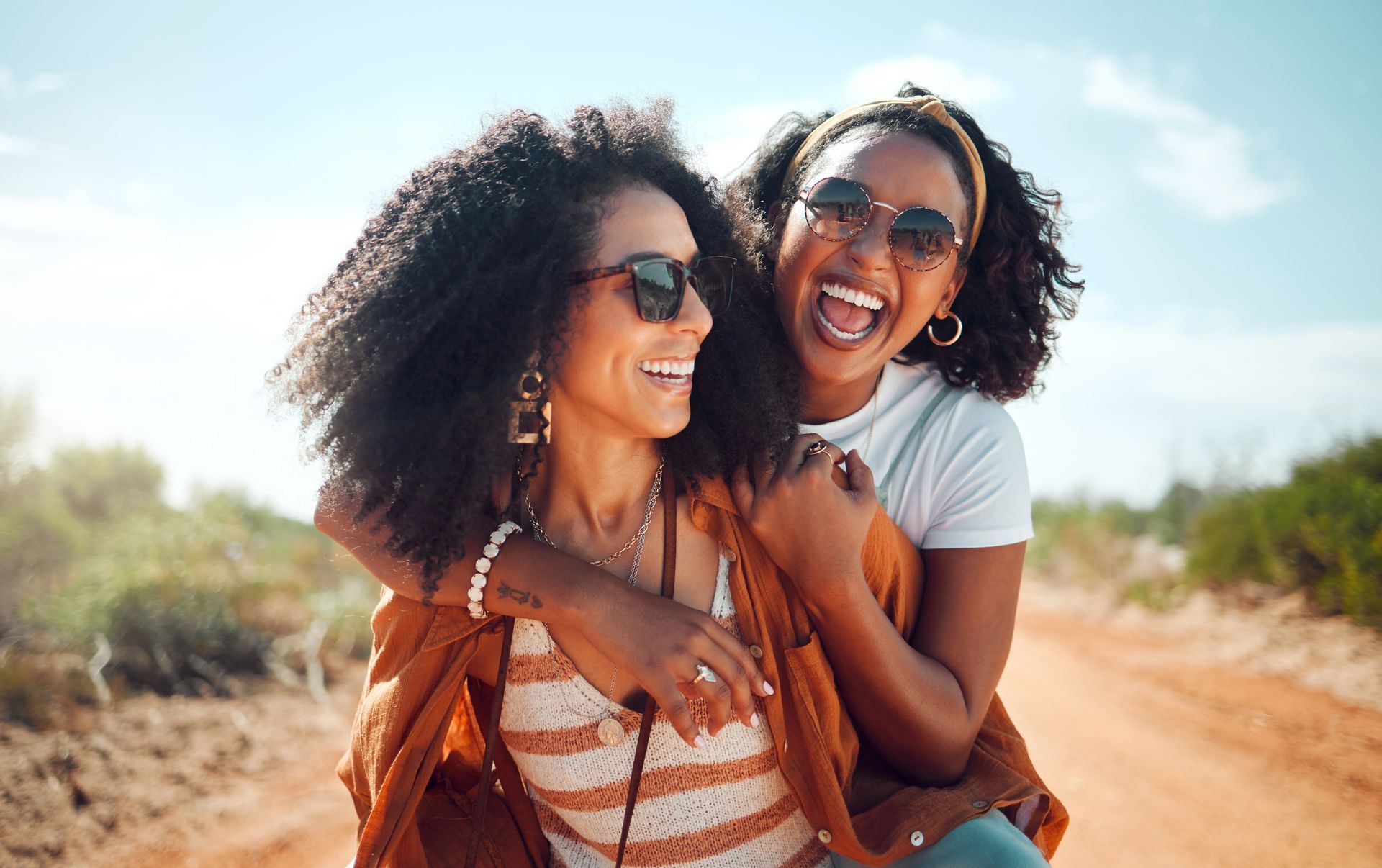 Two women are laughing and hugging each other on a dirt road.