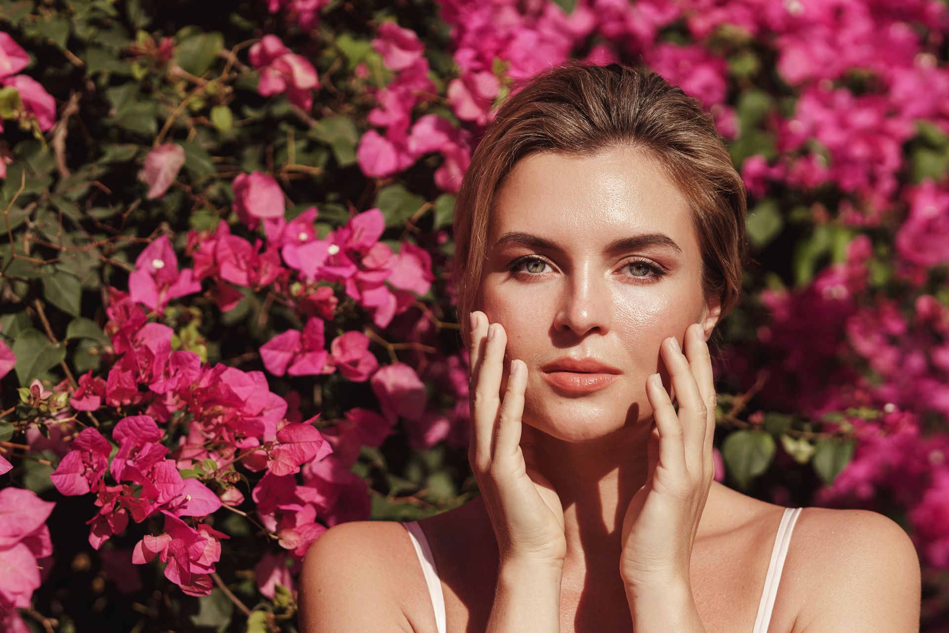 A person with their hands gently resting on their cheeks, posing in front of a vibrant bush of bright pink flowers.