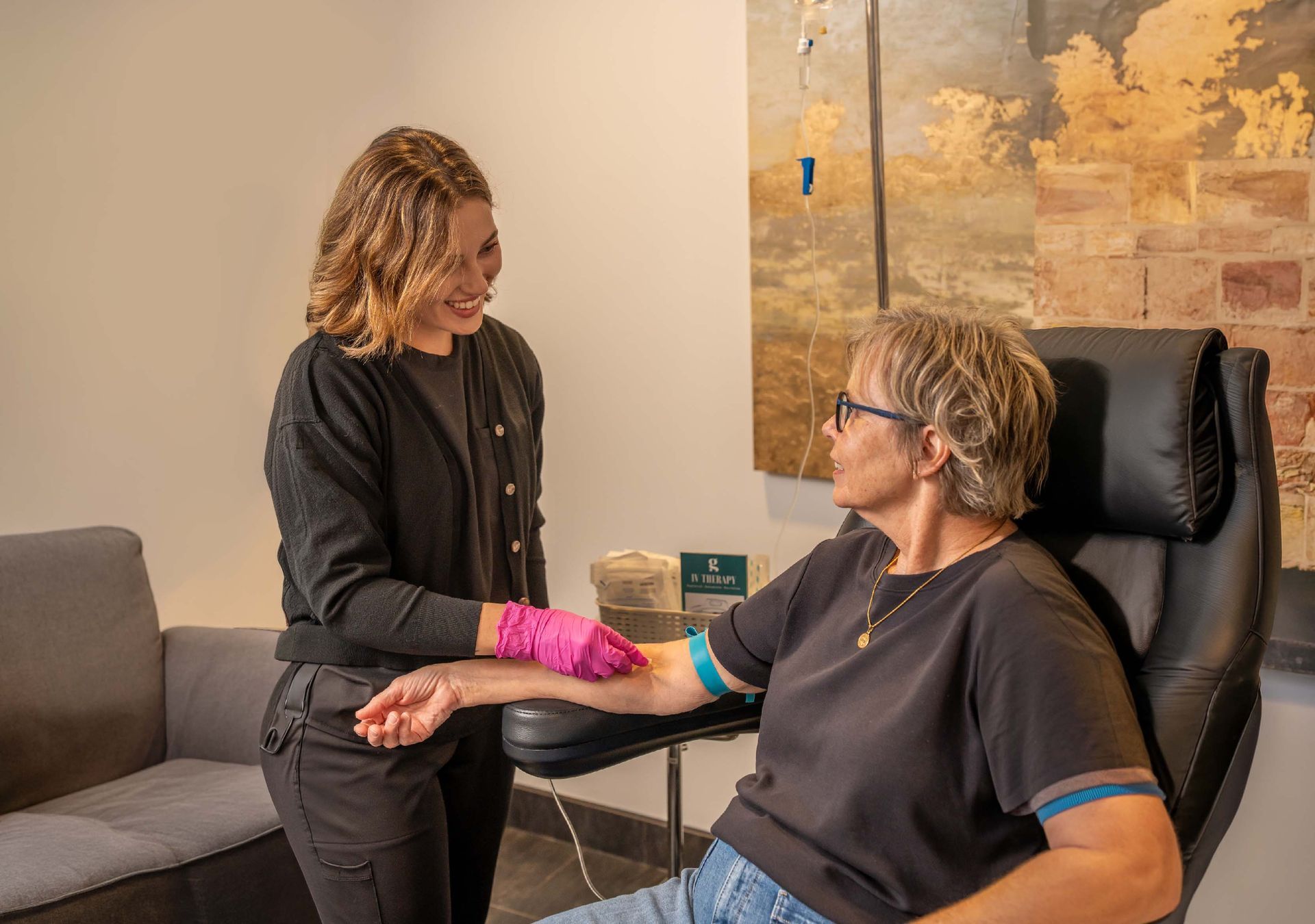 A blonde person in a white robe sits in a chair receiving an IV drip while reading a book in a white-tiled room.