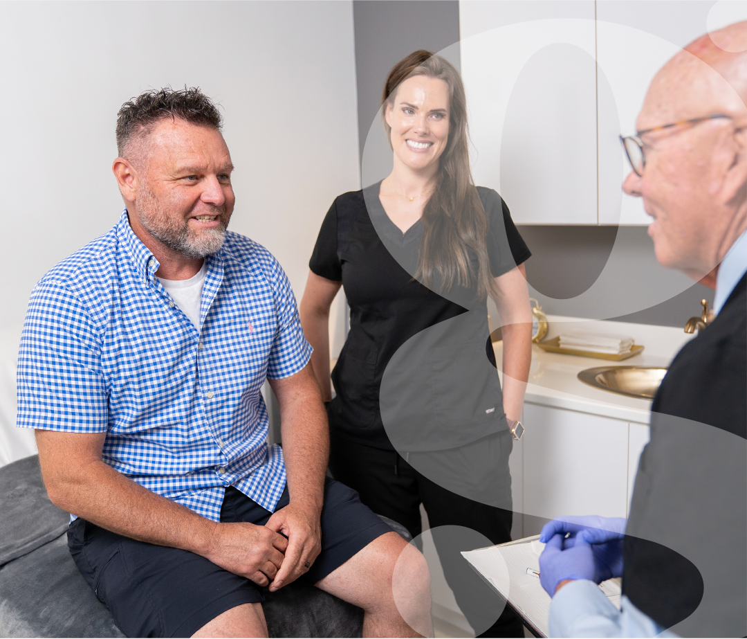 A man is sitting on a bed talking to a doctor while a woman stands behind him.