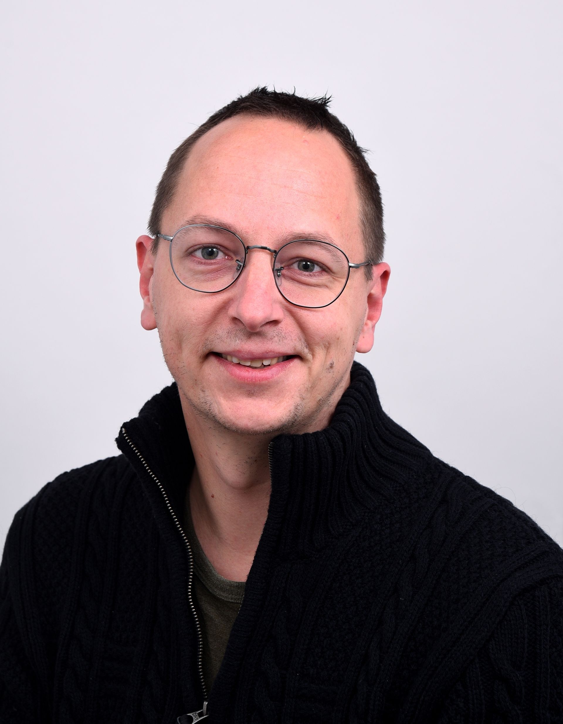 Een man met een bril en een zwarte trui lacht naar de camera