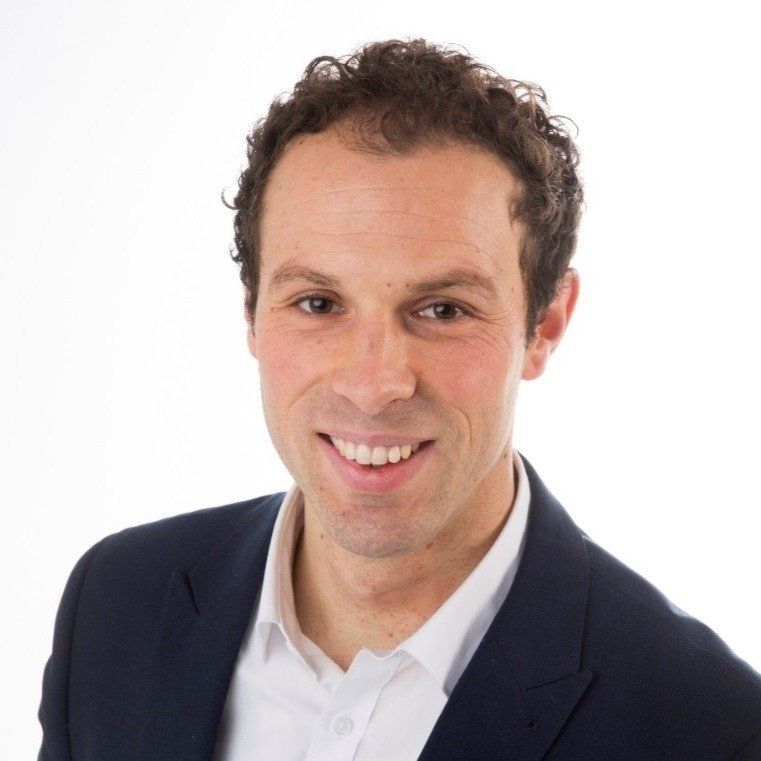 Een man in een pak en een wit overhemd lacht naar de camera