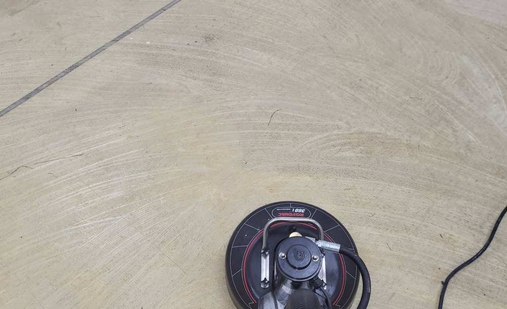 Floor buffer cleaning a light-colored, textured floor, with a black power cord attached.