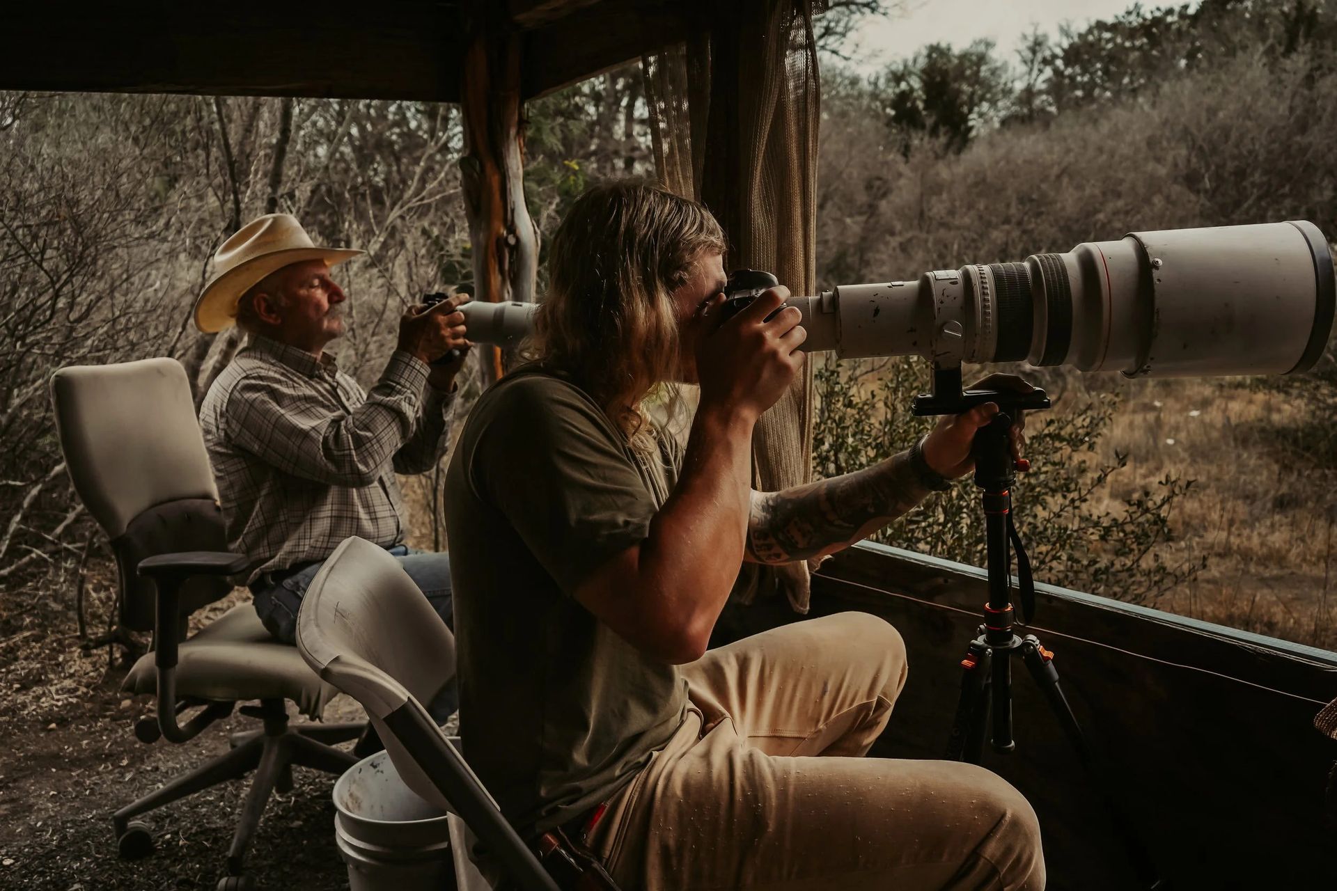 Two people using cameras with long lenses, seated in a wooden structure overlooking trees and bushes.