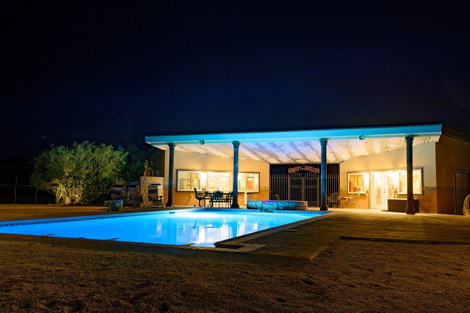 Night view of a building with a lit-up pool in front. Building has an awning and open windows.