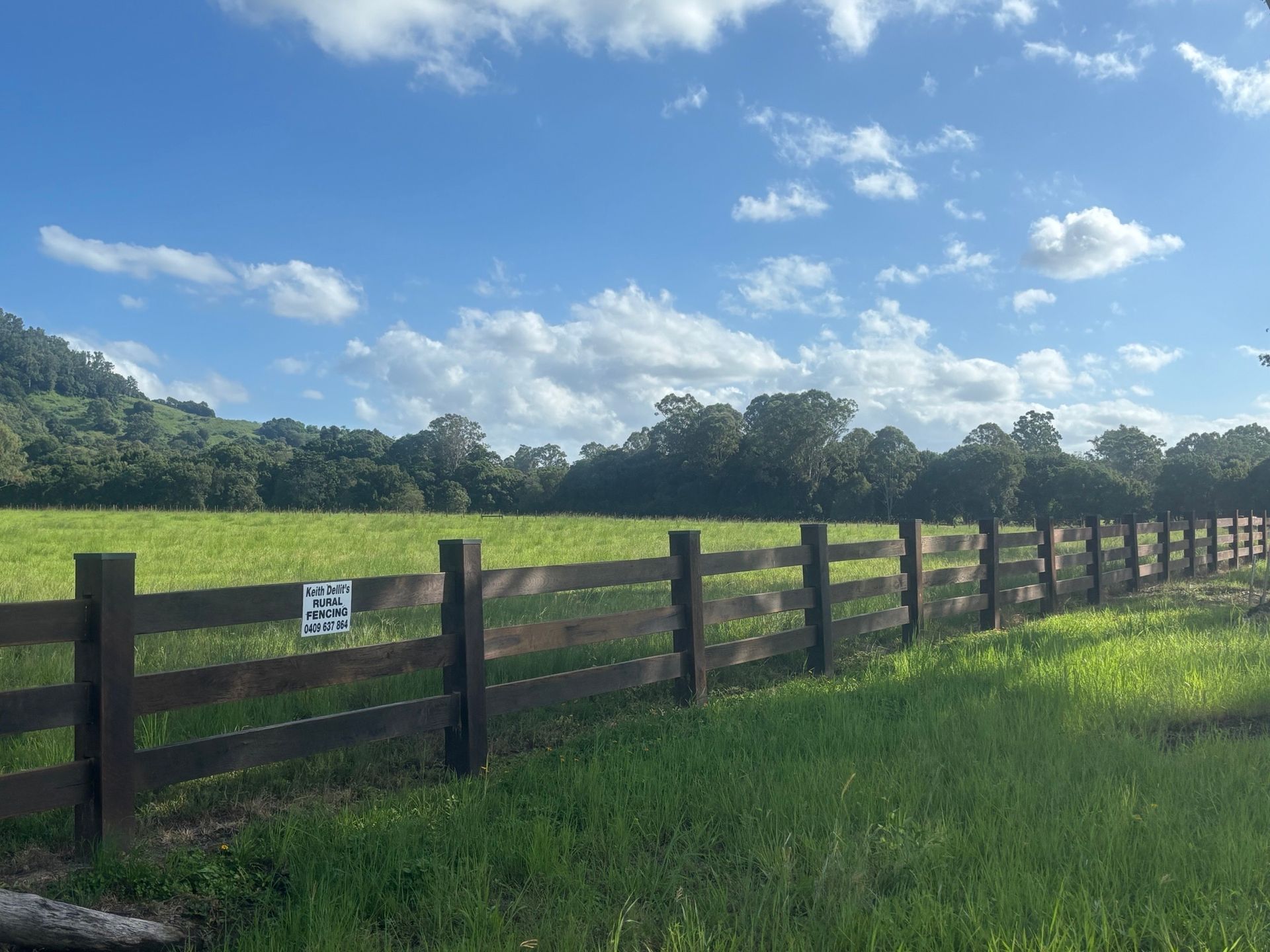 Green field bordered by a wooden fence under a partly cloudy blue sky; trees in the distance— Keith Dellit Rural Fencing in Yandina Creek, QLD