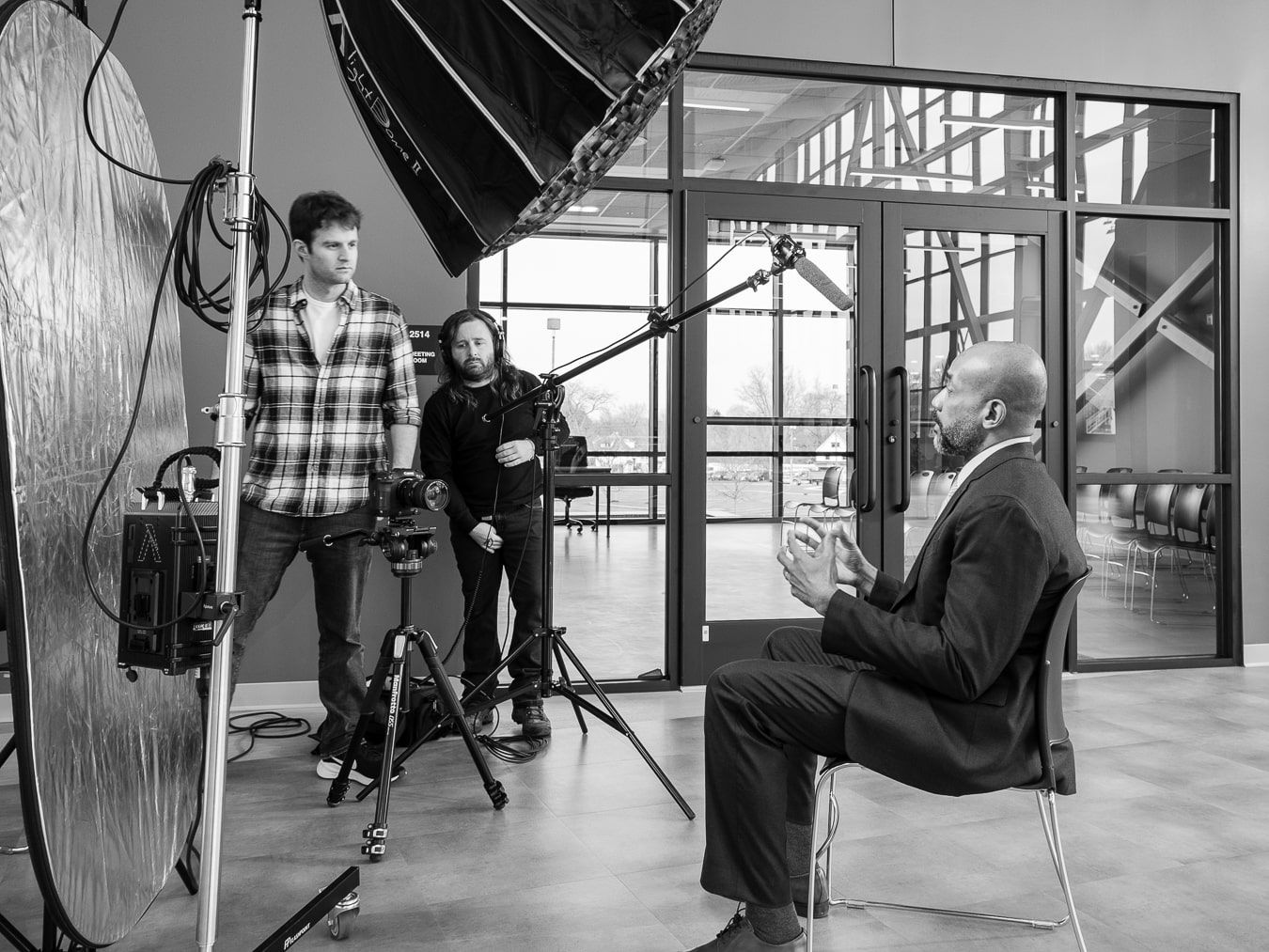 A man in a suit sits in front of a camera in a black and white photograph.