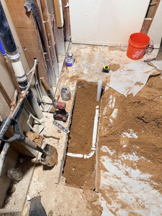 Bathroom renovation: plumbing pipes in a trench, red bucket, tools, and dirt.