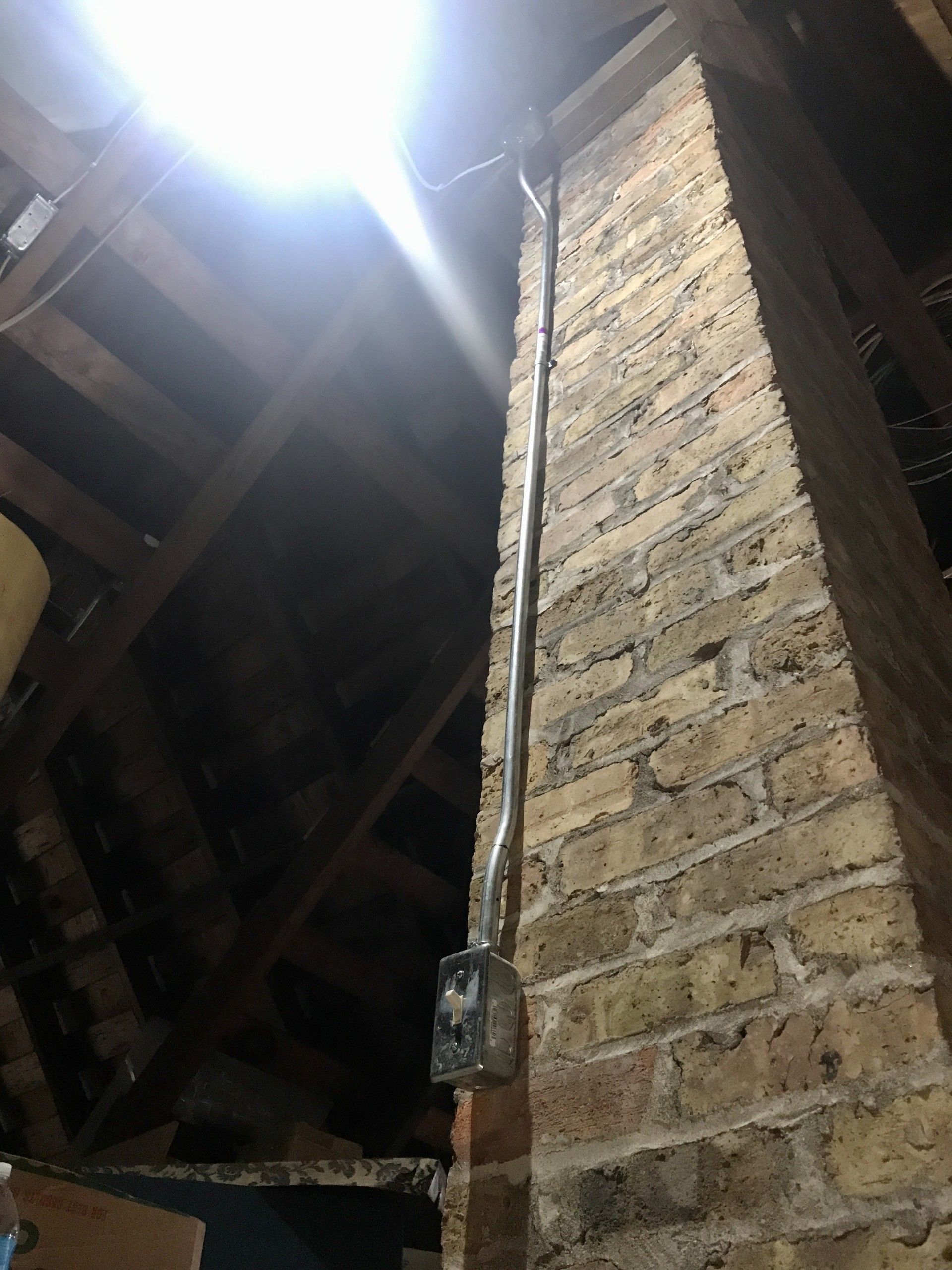 Electrical conduit and box attached to a brick chimney in an attic with wooden beams.