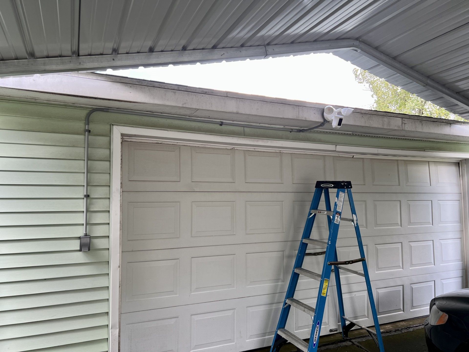 White garage door with blue ladder, under a carport, green siding on the left.