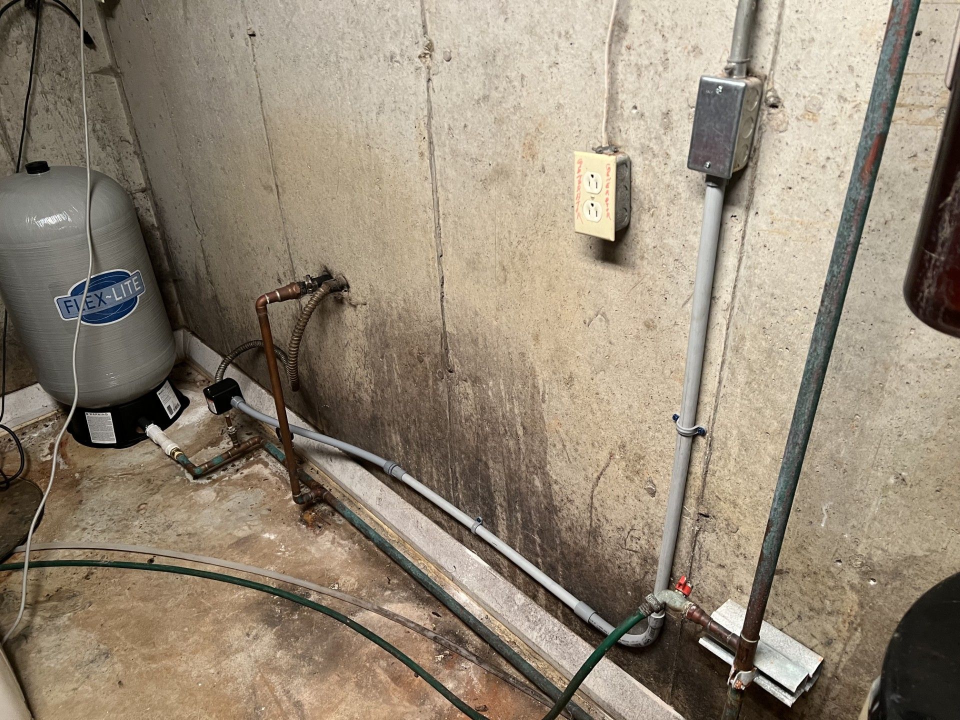 Water tank and pipes in a concrete basement, showing signs of water damage on wall.