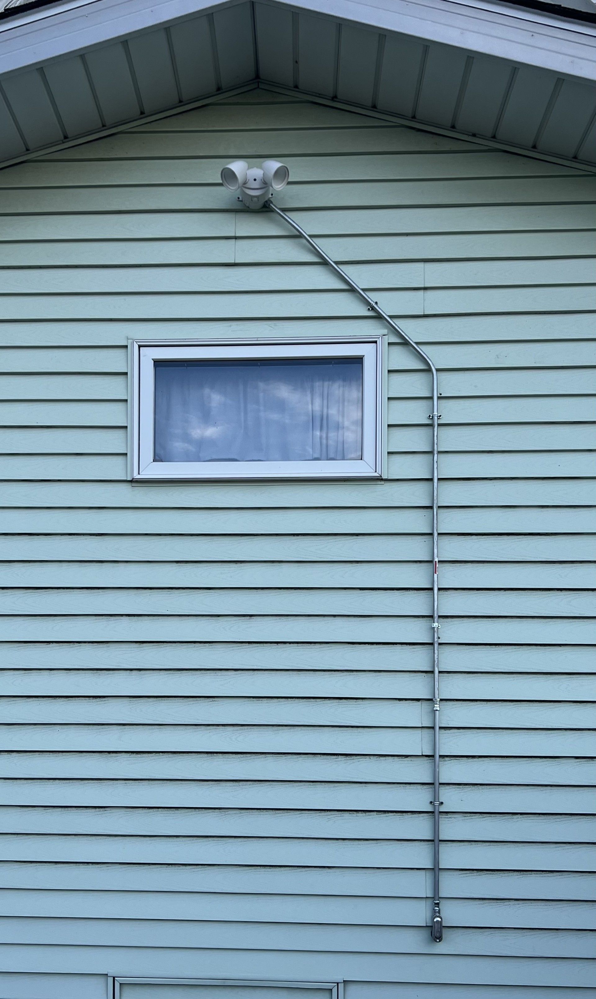 Light blue house siding with a window and a floodlight fixture connected by a metal conduit.