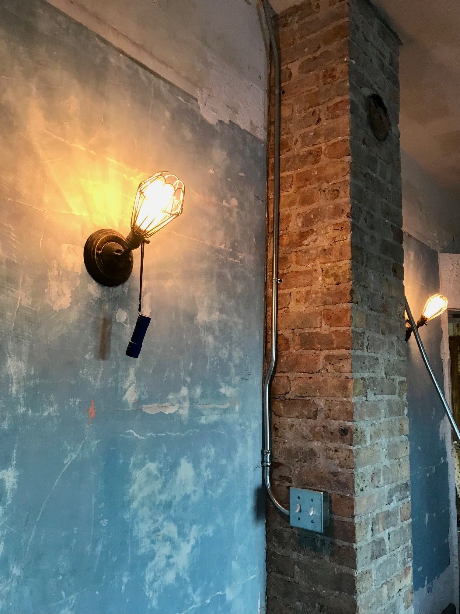Antique wall sconces on blue wall next to brick pillar, with exposed electrical conduit.