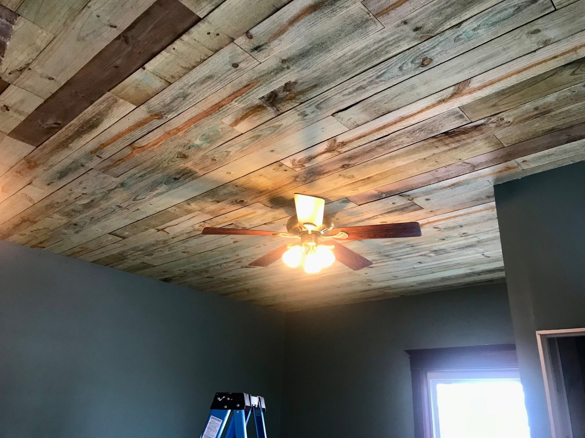 Ceiling covered in weathered wood planks with a ceiling fan. Dark green walls and window frame.