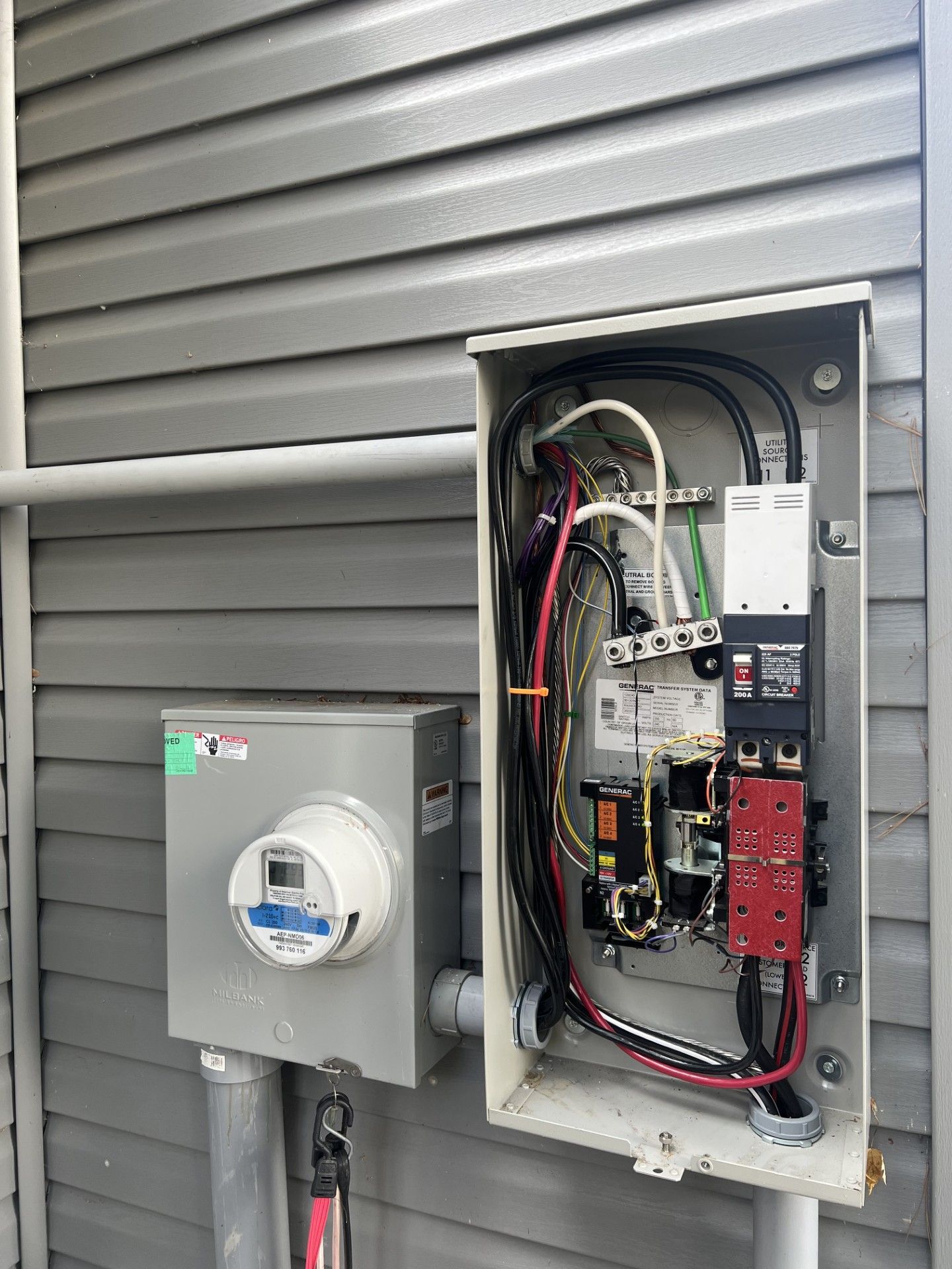 Electrical meter and panel mounted on a gray wall. Wiring and breakers visible inside the open panel.