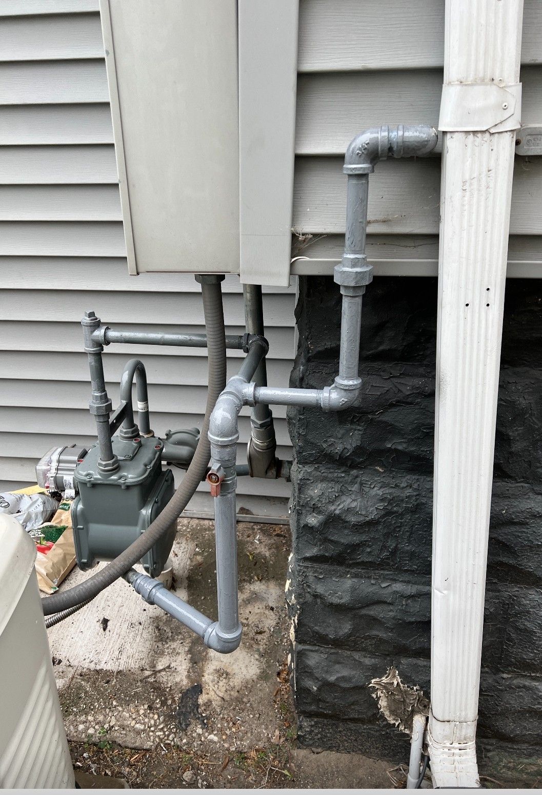 Gray gas meter and plumbing against a gray-sided building with a black stone foundation. White guttering on the right.