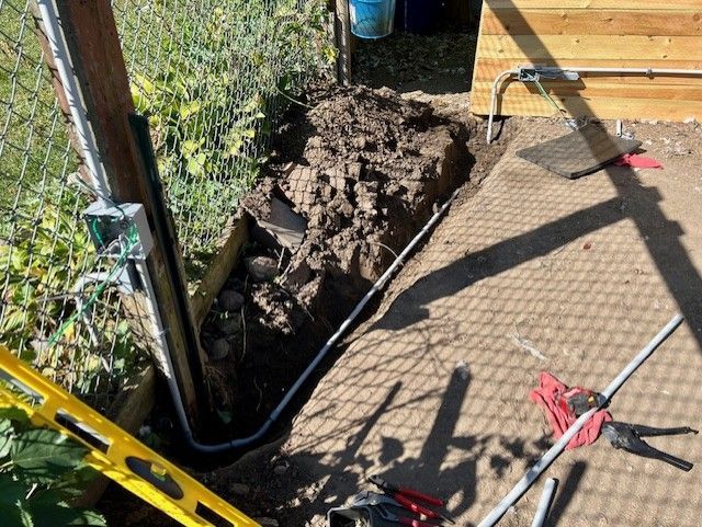 Trench dug in dirt next to concrete, with electrical conduit and tools present.