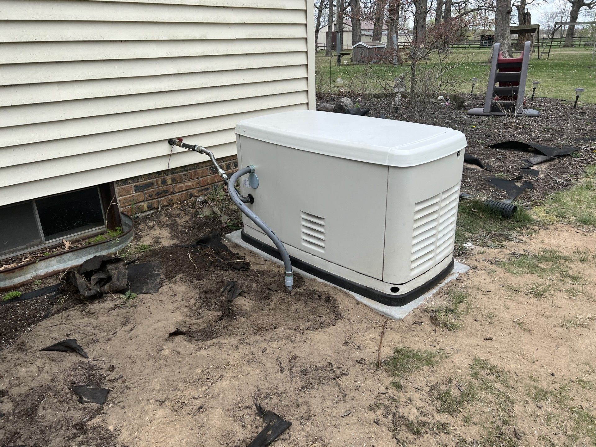 A beige generator sits beside a house on a concrete pad. A hose runs to it from the house's side.