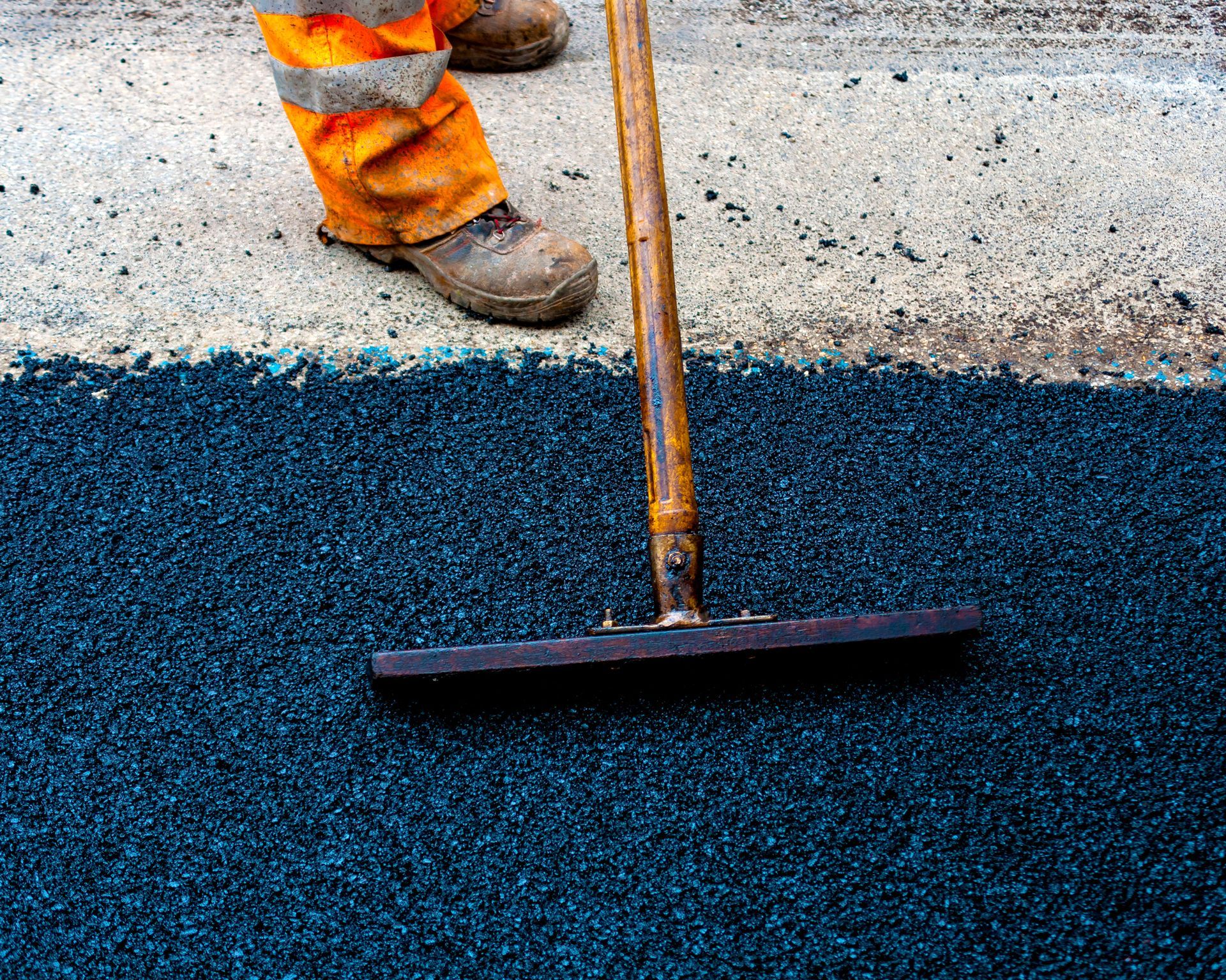 A worker rakes fresh black asphalt on a road surface, with orange safety pants visible.