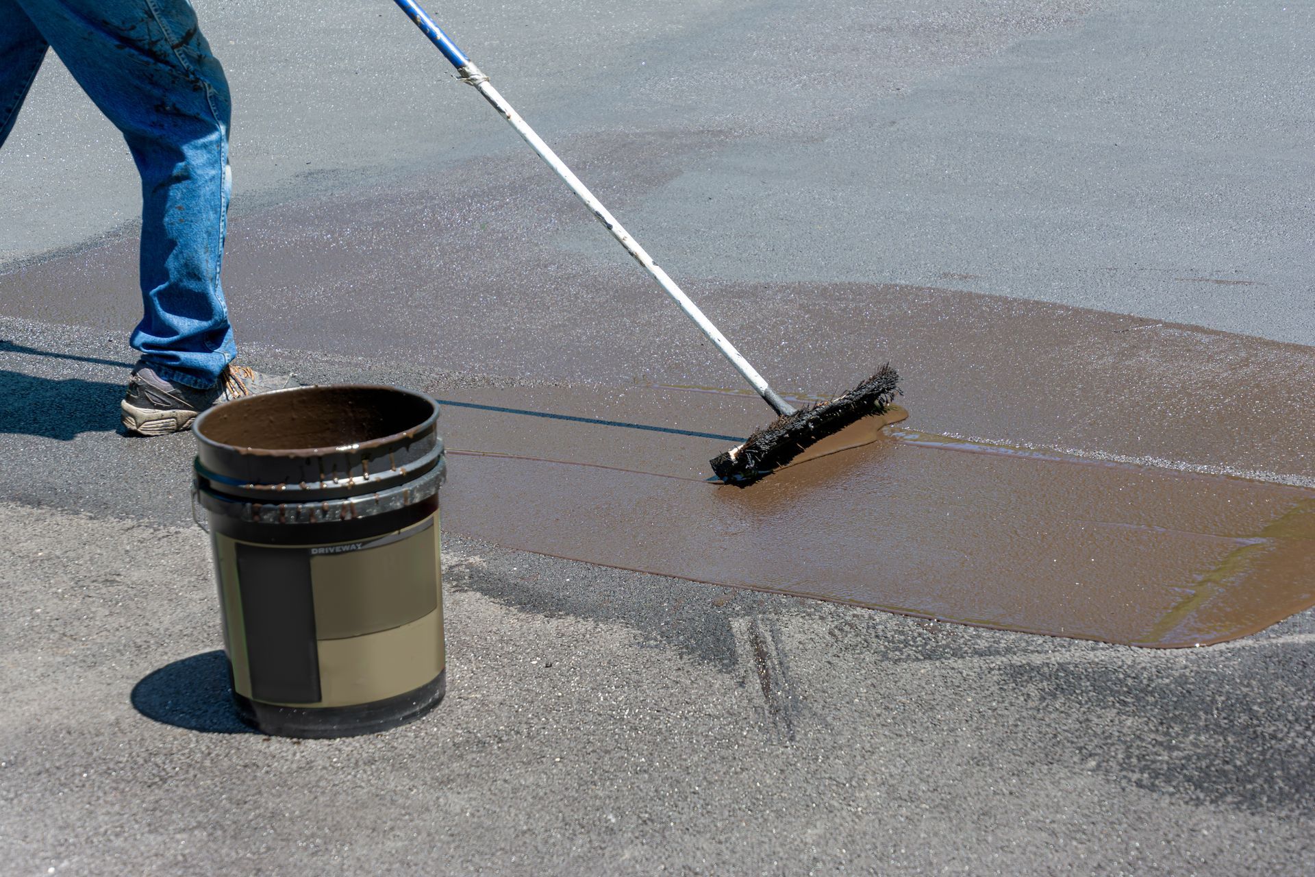 Person rolling black coating onto pavement beside a bucket on a sunny surface