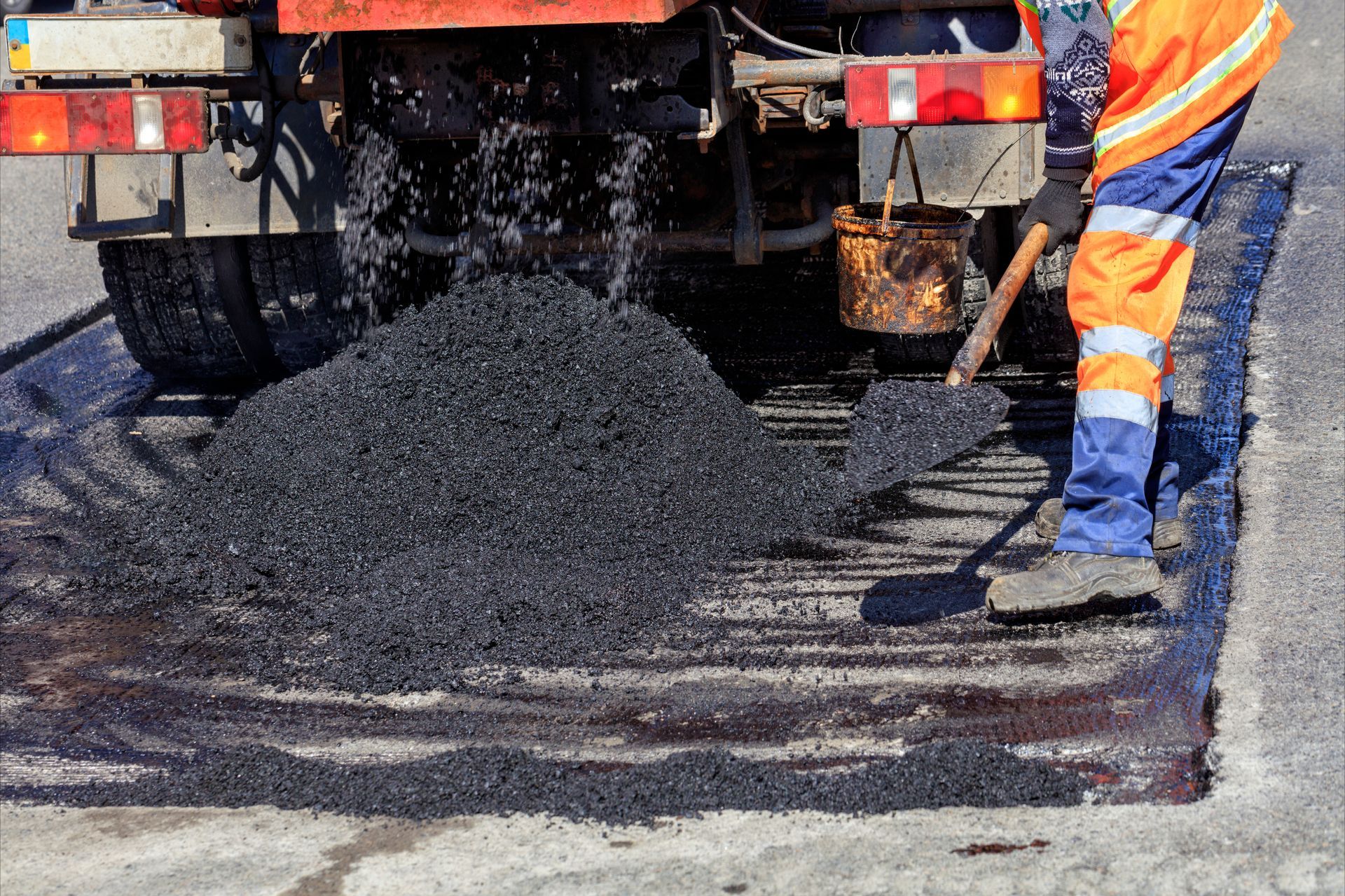 Road worker in orange safety gear spreading asphalt behind a truck, with a fresh black patch on the road.